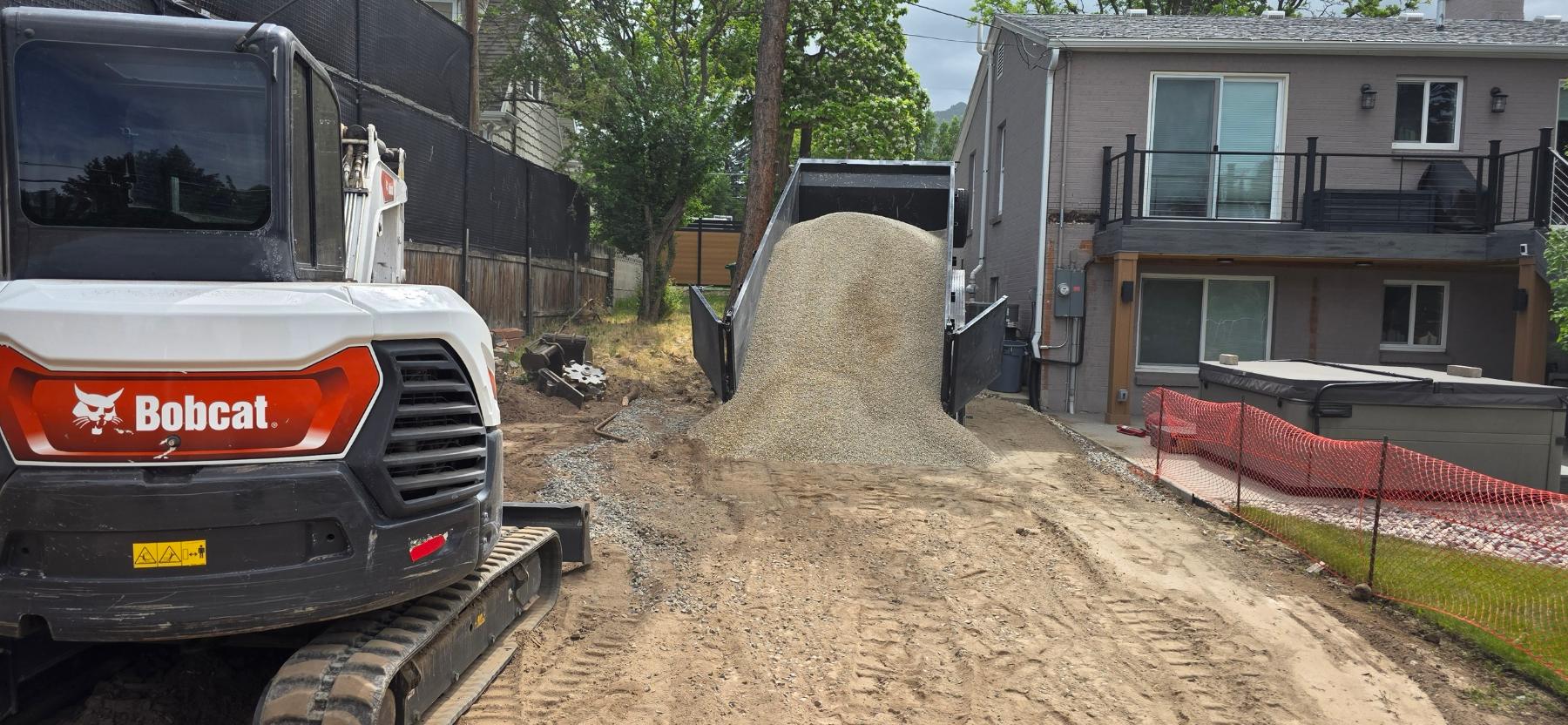 Road base being tipped into a narrow gap between two houses beside an excavator