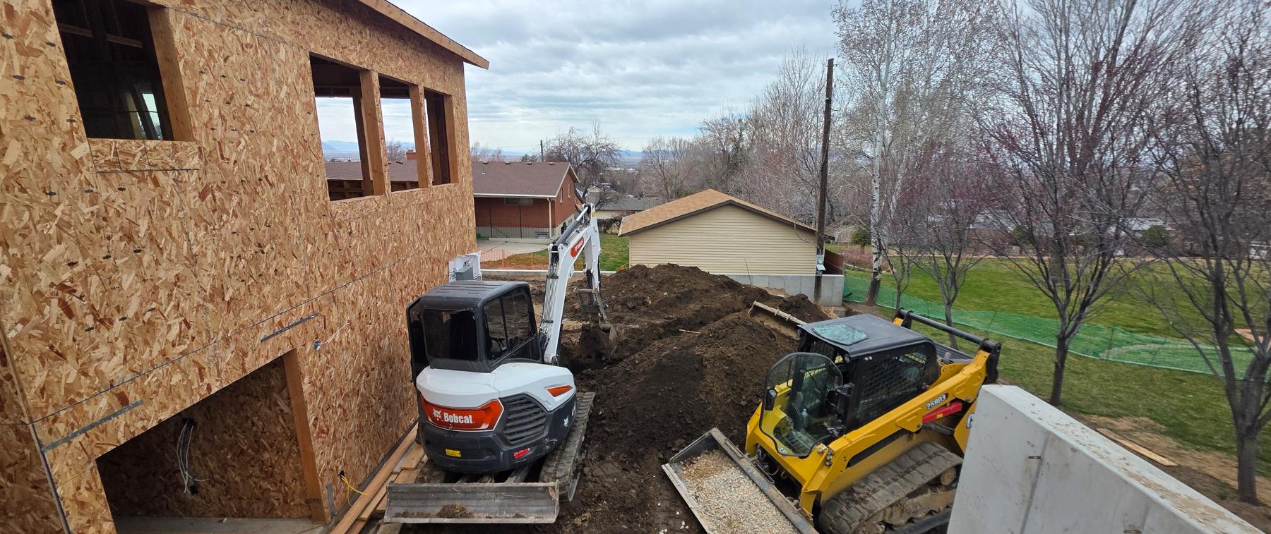 An excavator and a track loader backfilling against a newly framed two-story structure